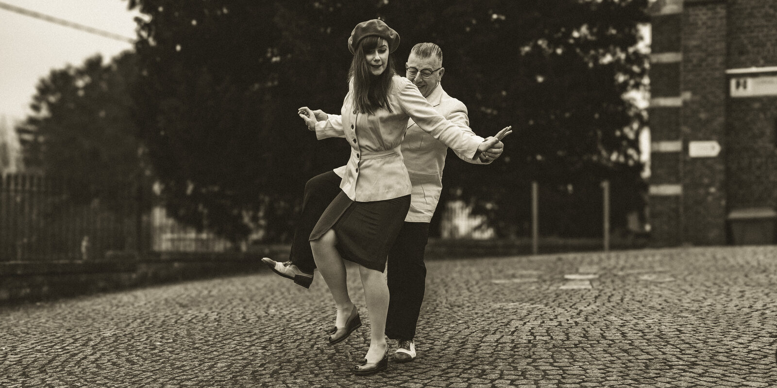 A couple in vintage-style clothing swing dancing on a cobblestone street, the man dipping his partner mid-step.