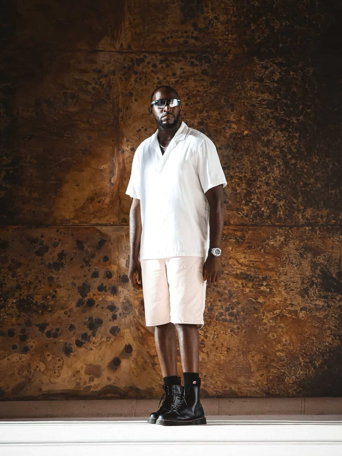 Choreographer Aurel Zola standing full-length in a white shirt, cream shorts and black boots against a weathered bronze wall, wearing angular sunglasses.