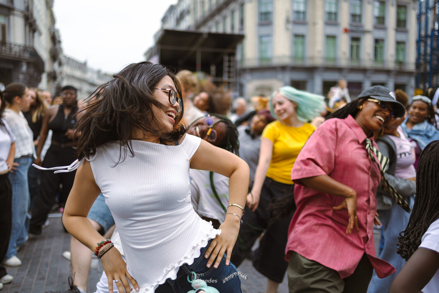A dancer in a white top, hair flying, laughing mid-move among a crowd dancing in a Brussels street with an open-air stage behind them