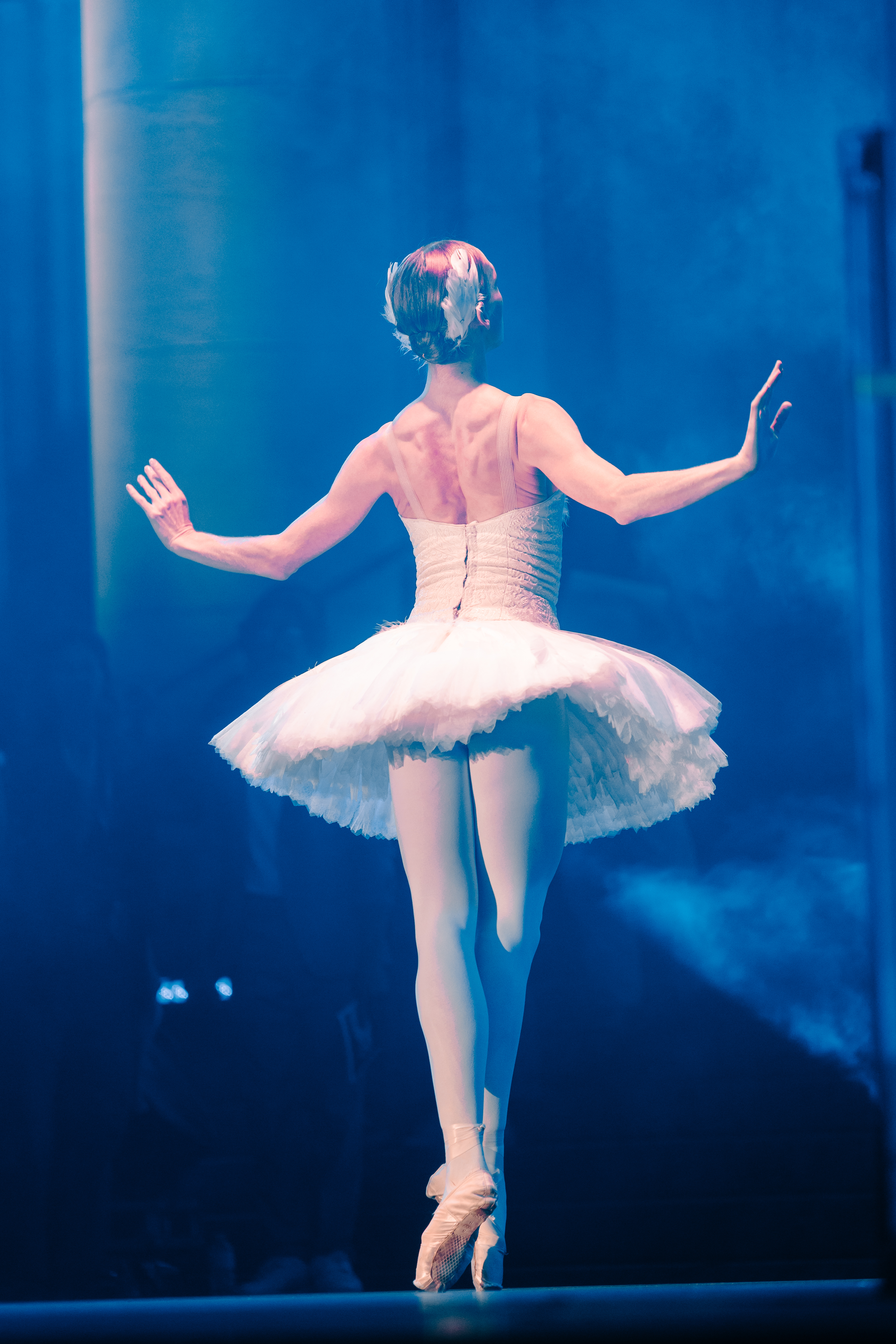 Ballet dancer in a white tutu photographed from behind, balancing en pointe under blue stage spotlights