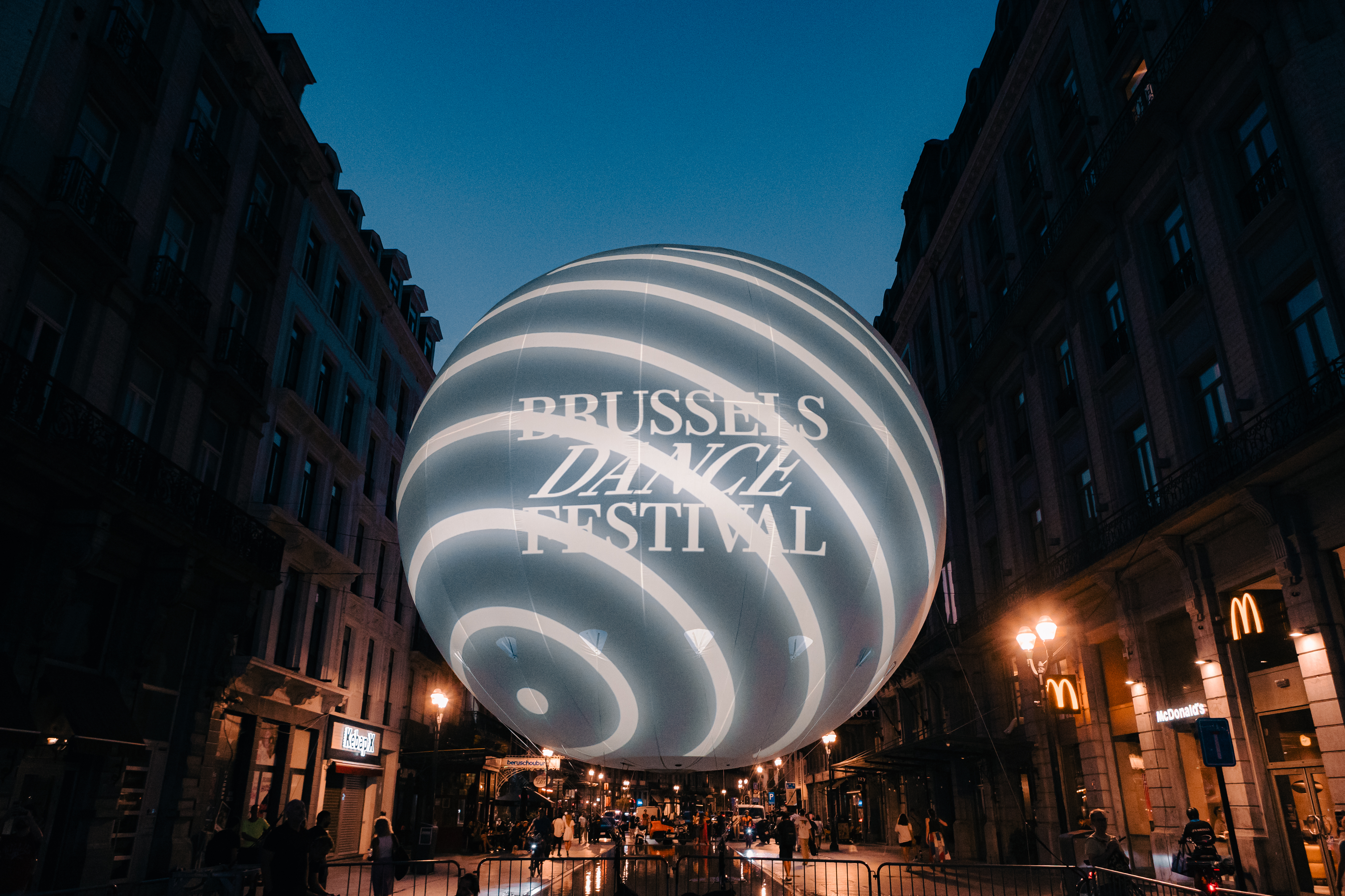 The giant illuminated Brussels Dance Festival globe floating between the facades of Brussels city centre at dusk, with 'Brussels Dance Festival' text projected on its surface