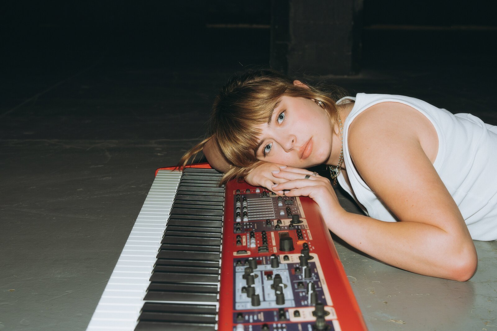 Billie du Page lying on a dark studio floor in a white sleeveless top, resting her head and folded hands on a red stage keyboard and looking straight at the camera