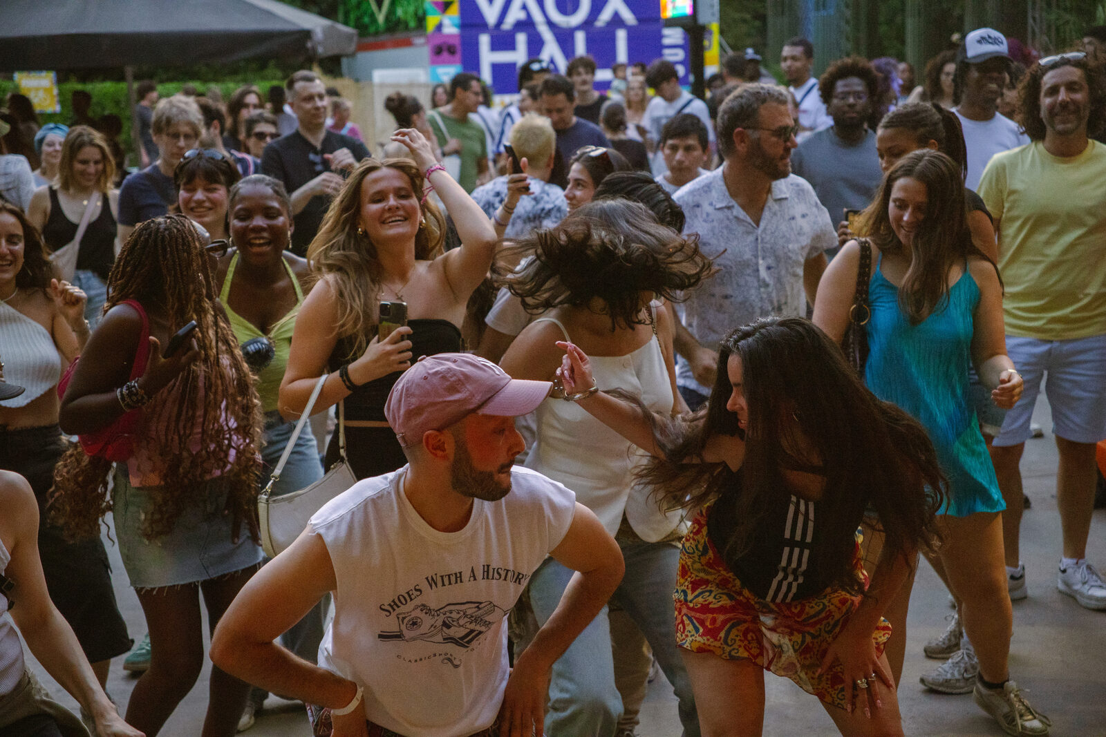 A large crowd dancing together outdoors near a 'Vaux Hall' venue sign at golden hour.