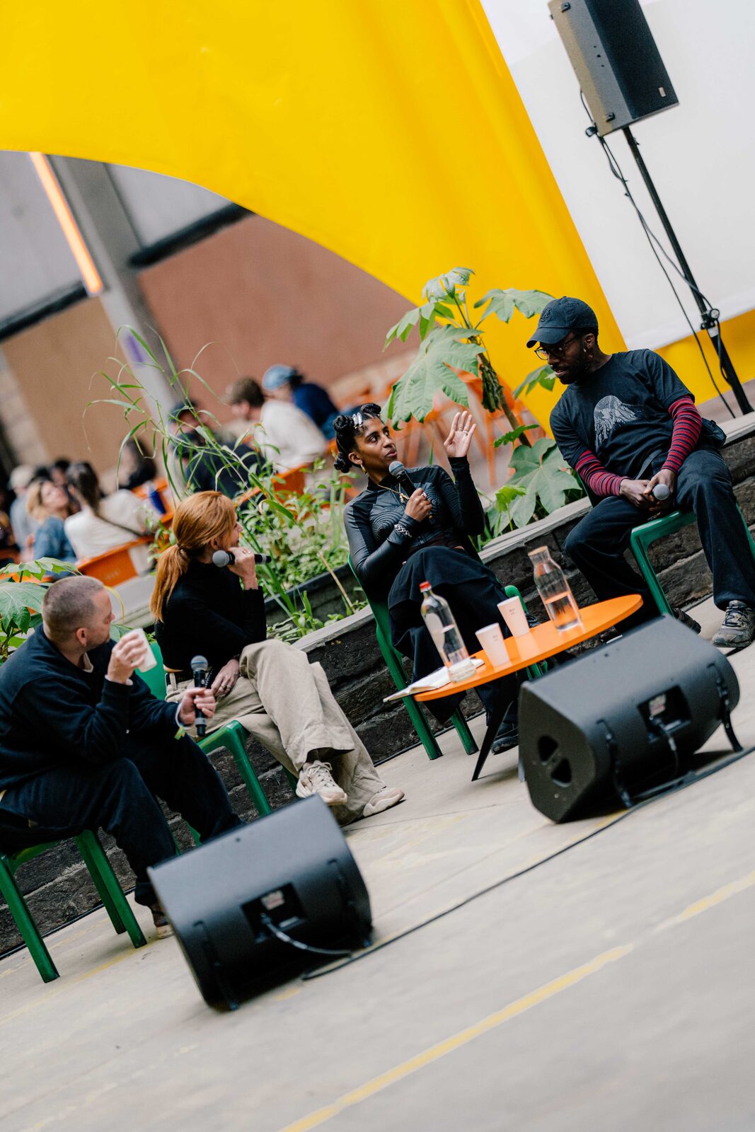 Panel speakers holding microphones during an outdoor discussion, seated among plants under a yellow canopy.