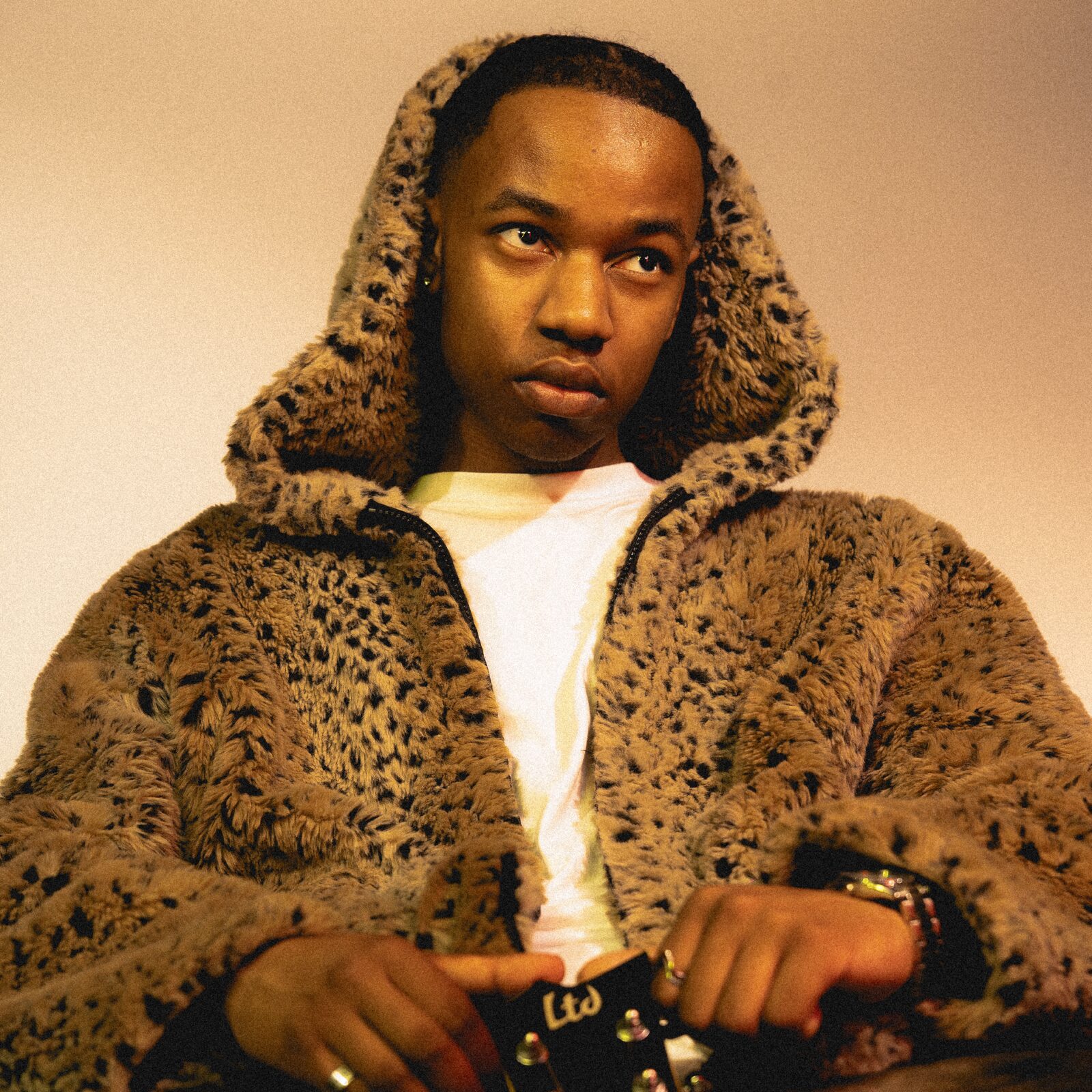 Portrait of a performer in a leopard-print faux-fur hooded coat over a white T-shirt, hands resting on a studded belt, looking to the side against a warm beige backdrop