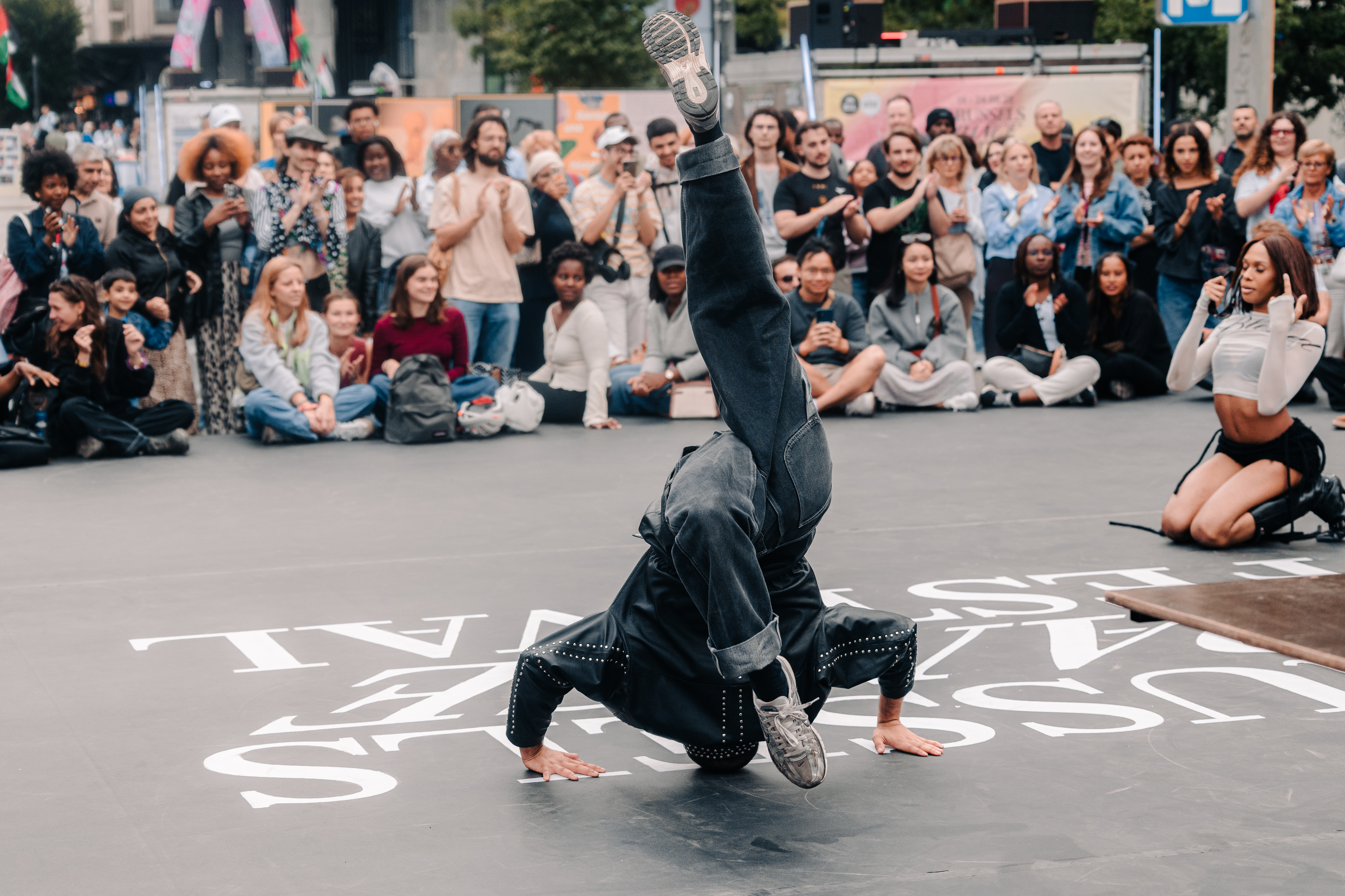 A breakdancer freezing on one hand mid-headspin on the Place De Brouckère stage floor, painted with the Brussels Dance Festival wordmark, surrounded by a seated crowd of spectators