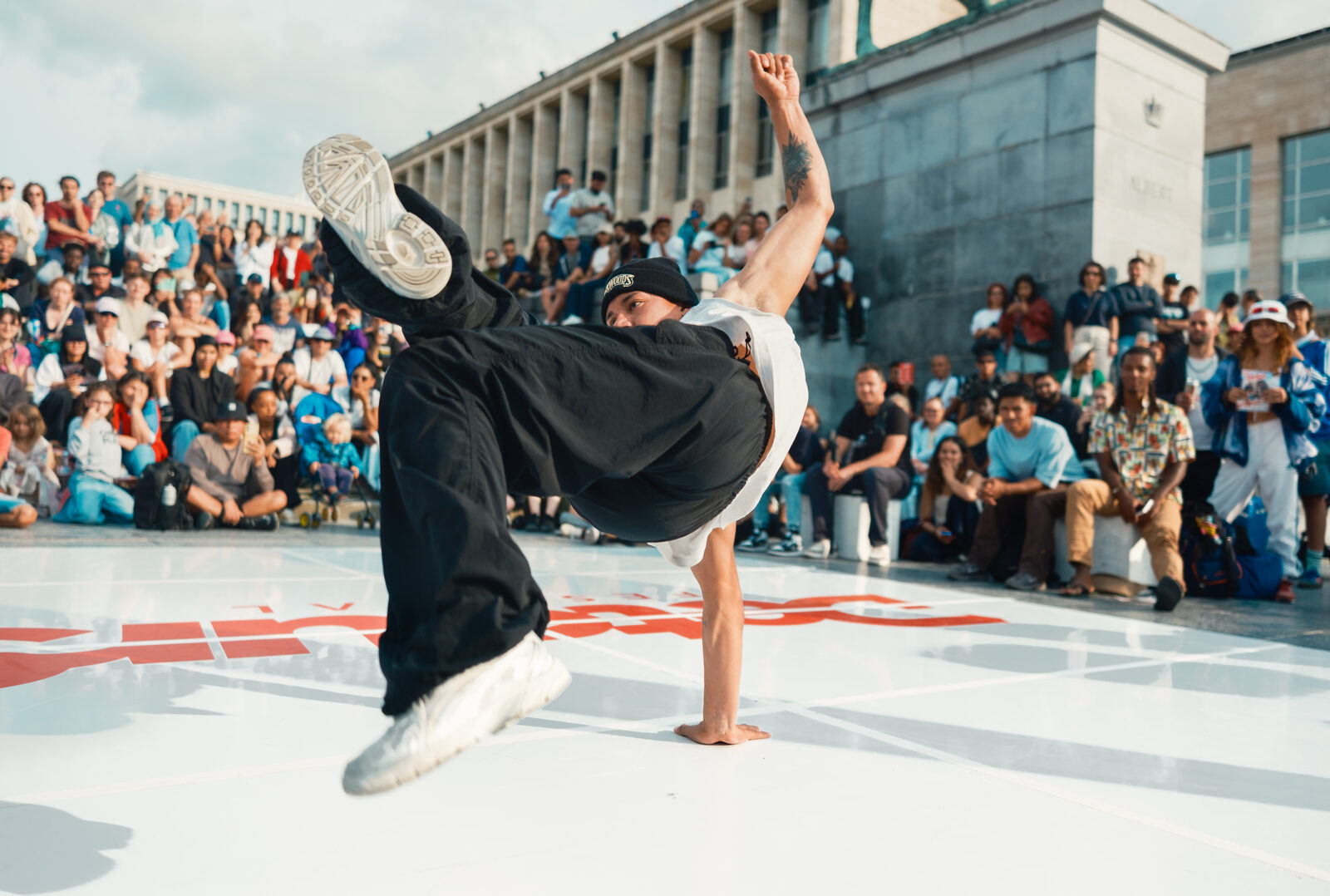 A breakdancer performing a one-handed freeze on an outdoor stage, watched by a large seated crowd in front of a monumental building.