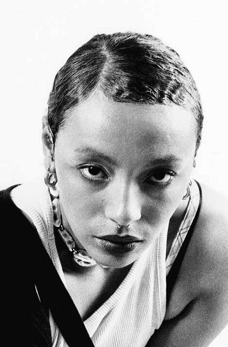 Black-and-white close-up portrait of a DJ with finger-waved cropped hair, hoop earrings and a chain necklace, leaning towards the camera in a white ribbed vest