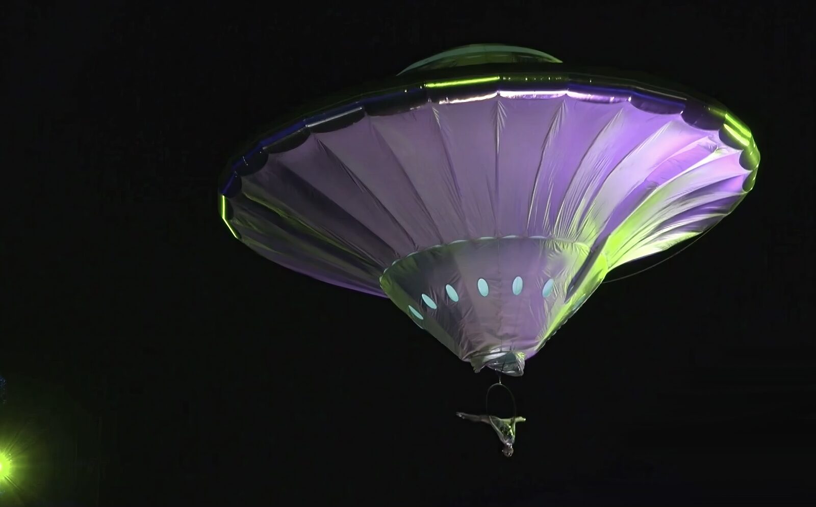 A giant illuminated purple and green balloon shaped like a flying saucer, with an aerialist performing on a hoop suspended beneath it against the night sky.