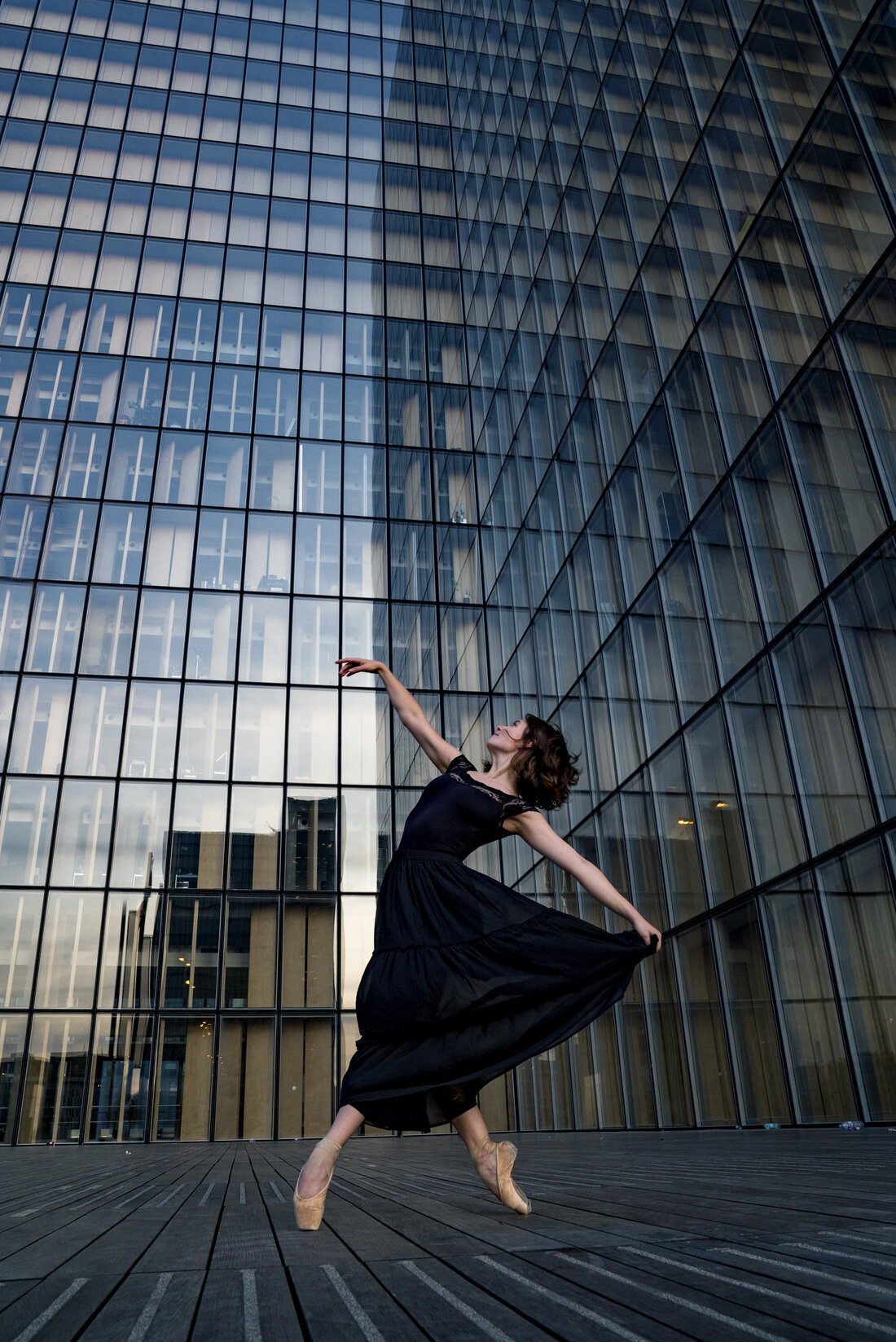 A ballerina in a black dress dancing en pointe against the glass facade of a modern office tower.