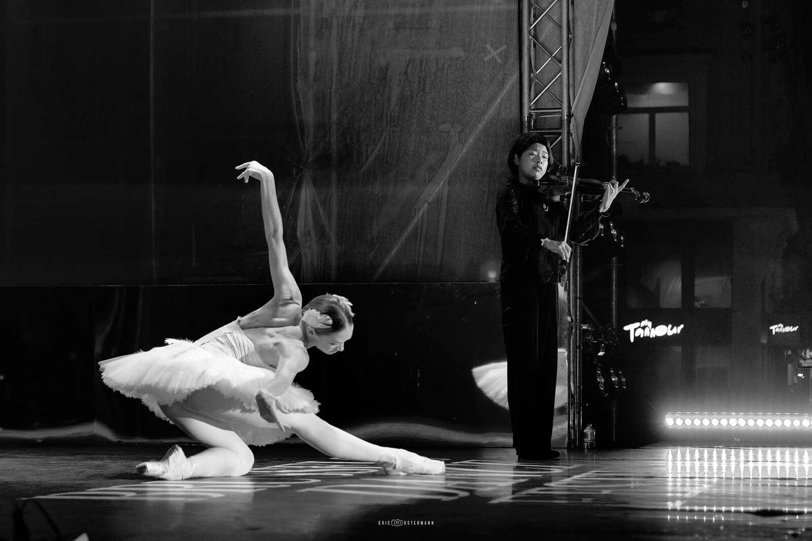 A ballerina in a white tutu balancing en pointe beside a violinist playing on an outdoor stage at dusk.