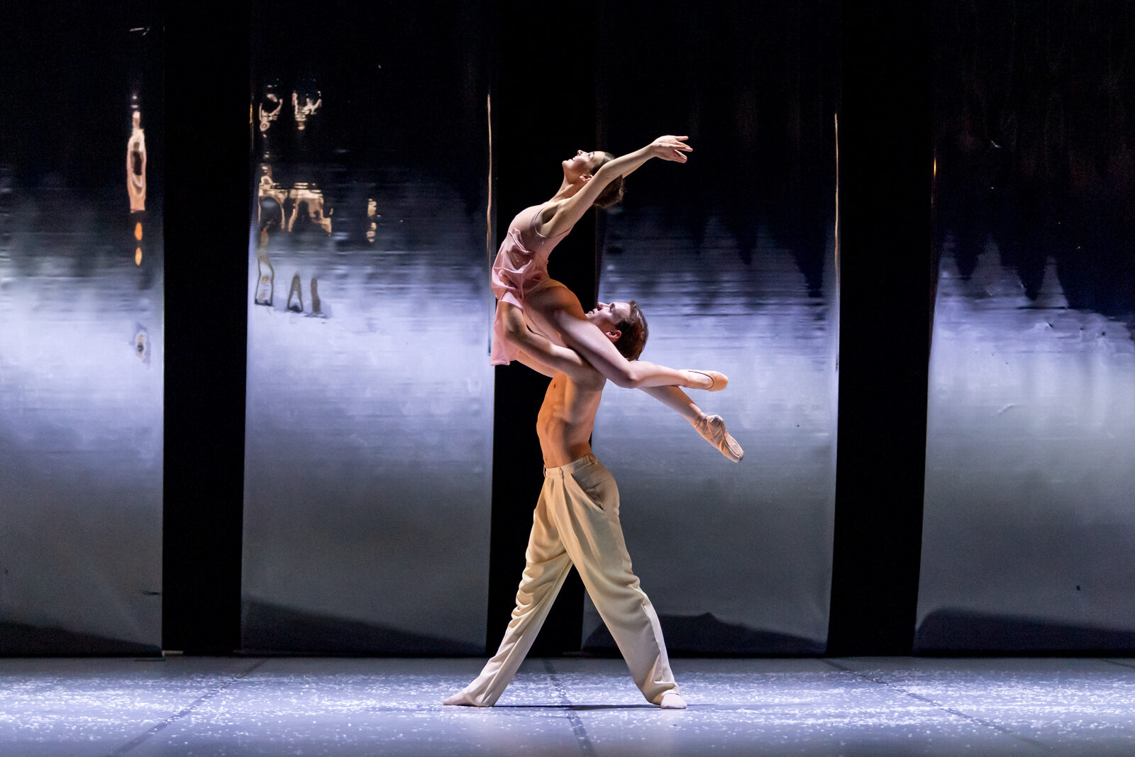 Two ballet dancers on a dark stage: a bare-chested dancer in cream trousers lifts a partner in a pink leotard who arches backwards with one arm outstretched, in front of reflective metallic panels