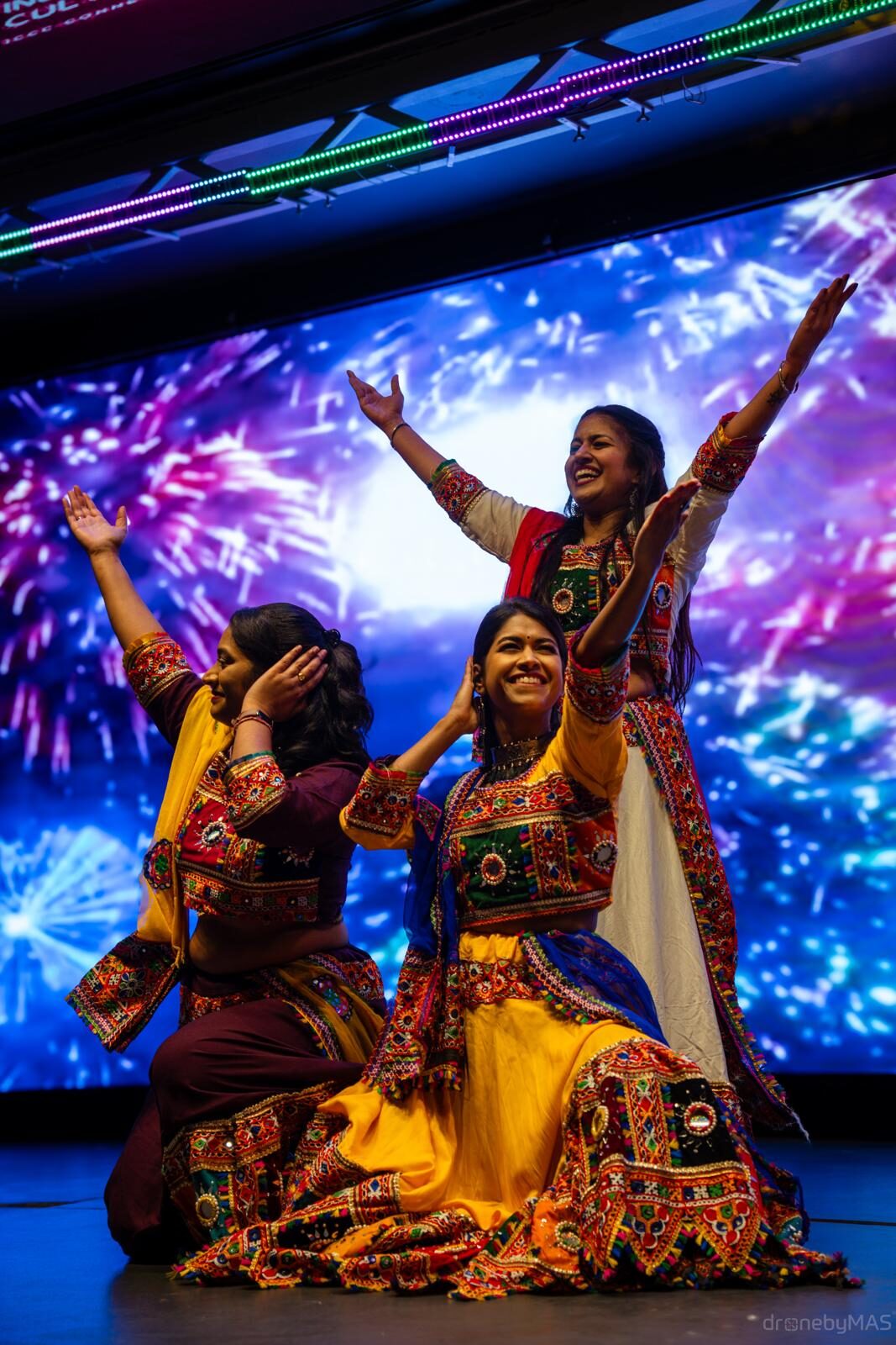 Three dancers in colourful, richly embroidered traditional Indian costumes performing on stage with arms raised, a fireworks projection lighting the backdrop.