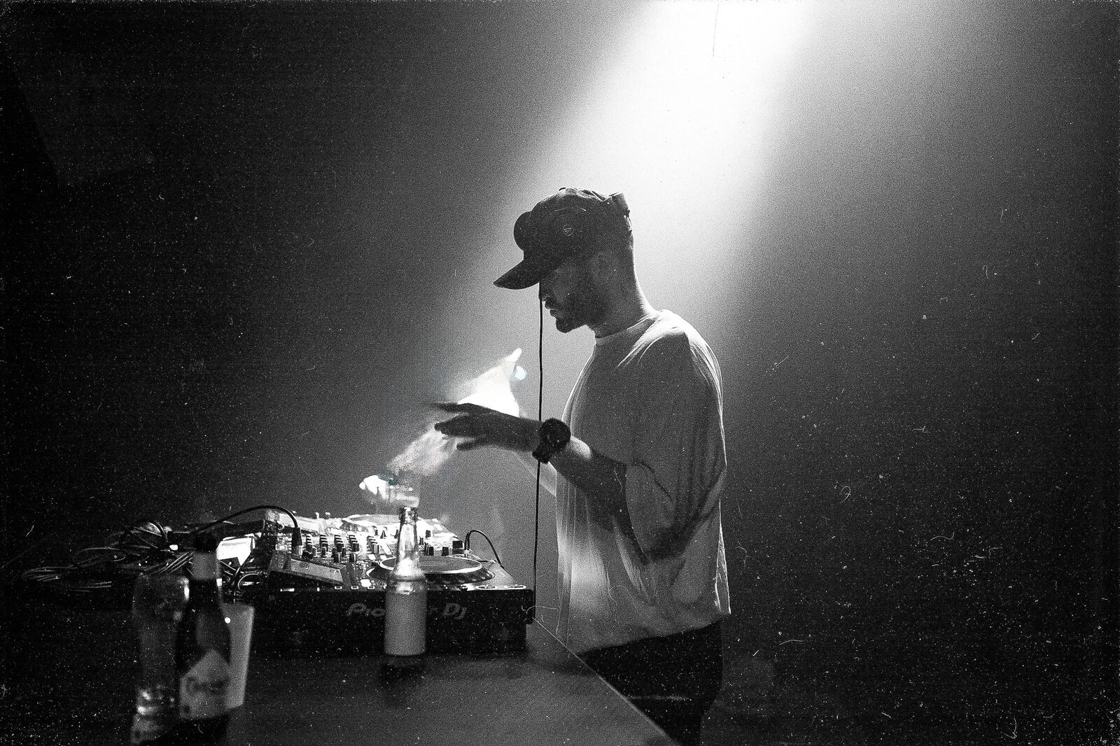 Black-and-white photo of a DJ in a cap working a Pioneer deck, lit by a single beam of light in a dark room
