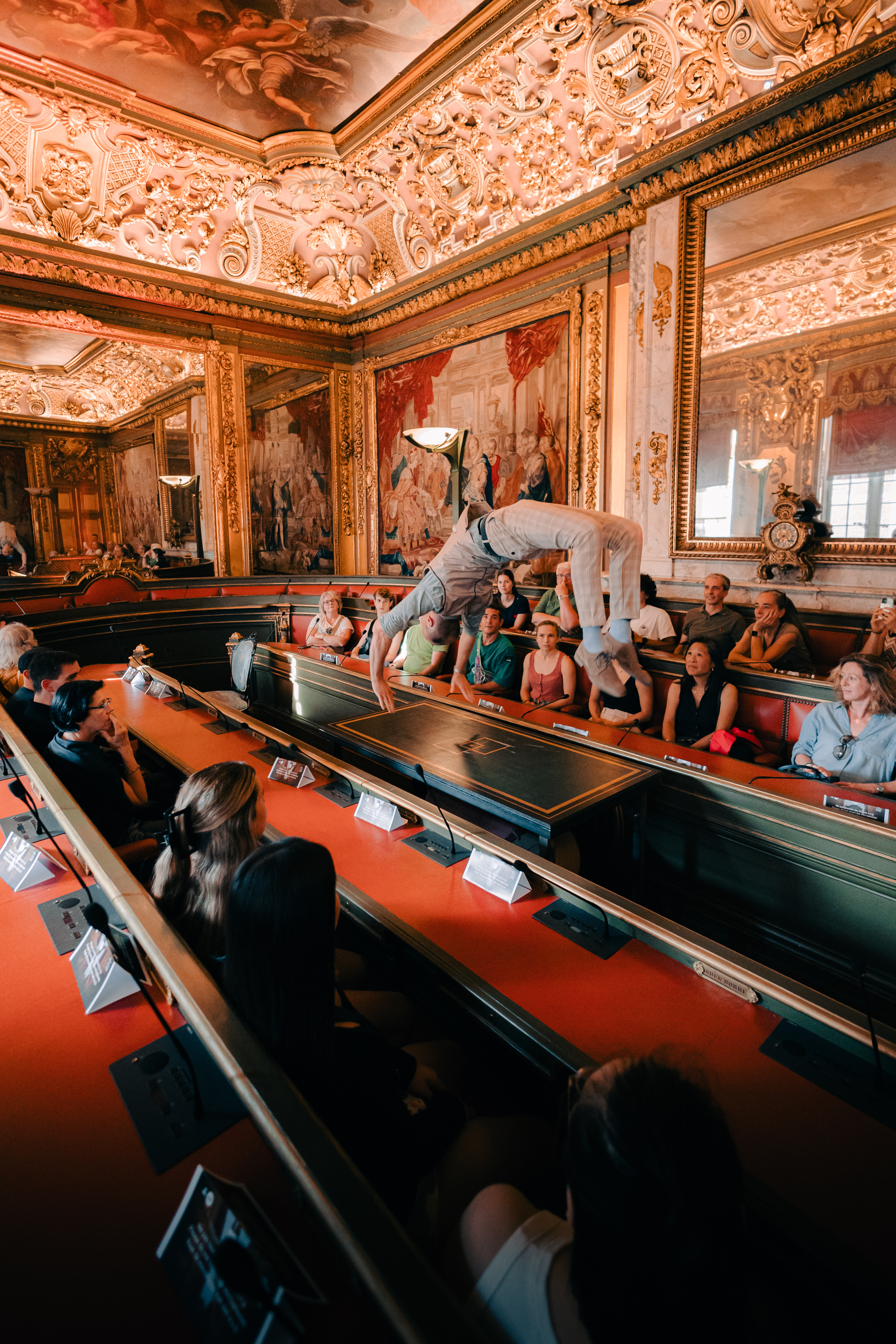 A dancer arching into a backbend on a table inside the Hôtel de Ville's gilded council chamber, watched by an audience seated along the room's red-upholstered benches under an ornate painted ceiling