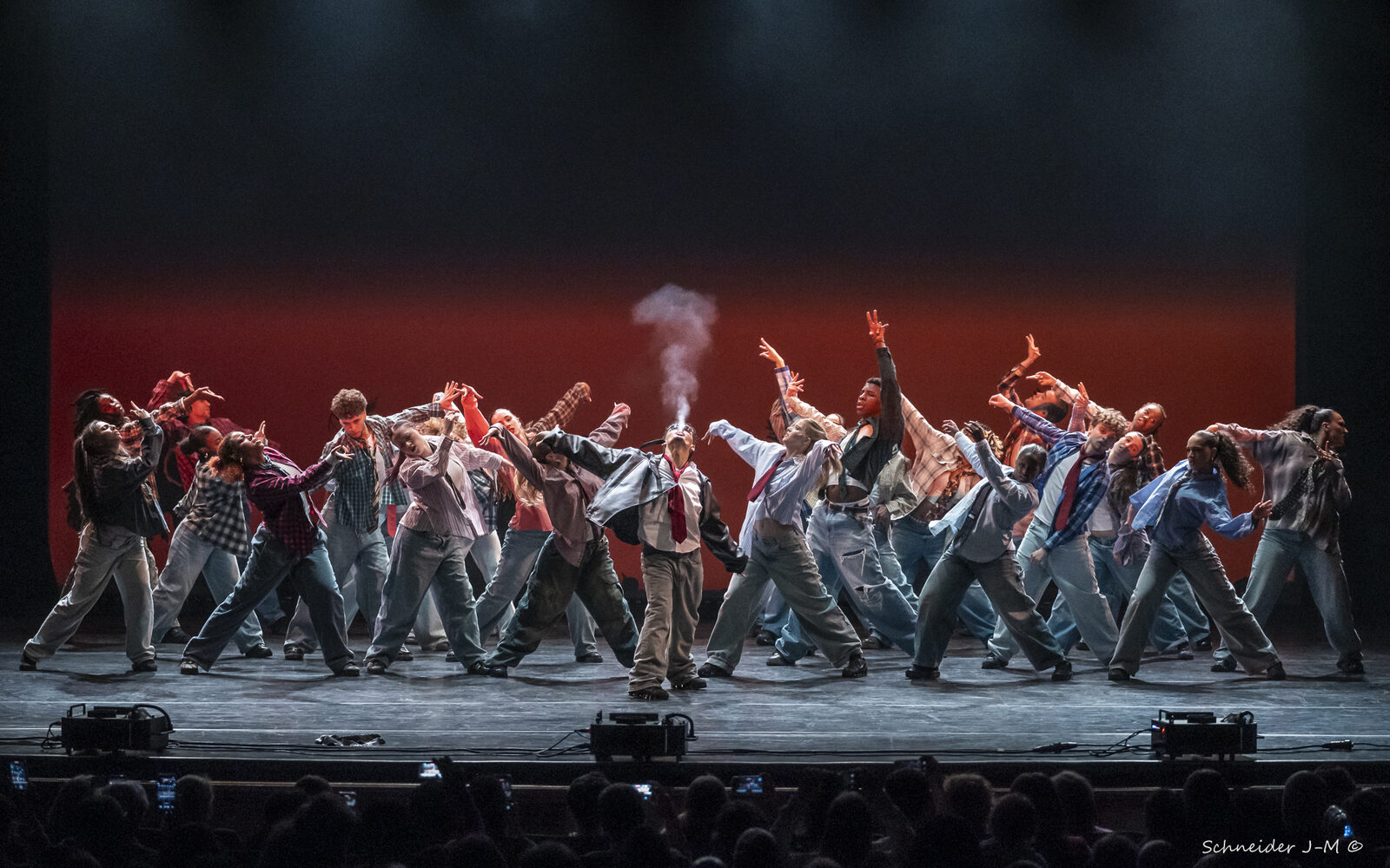 A large ensemble of dancers lunging in unison with arms raised on a theatre stage lit in deep red, with silhouettes of the audience in the foreground