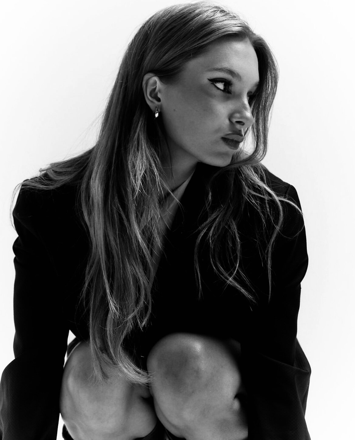 Black-and-white studio portrait of singer Noée crouching in an oversized dark blazer, long hair falling forward as she looks off to the side against a plain pale backdrop