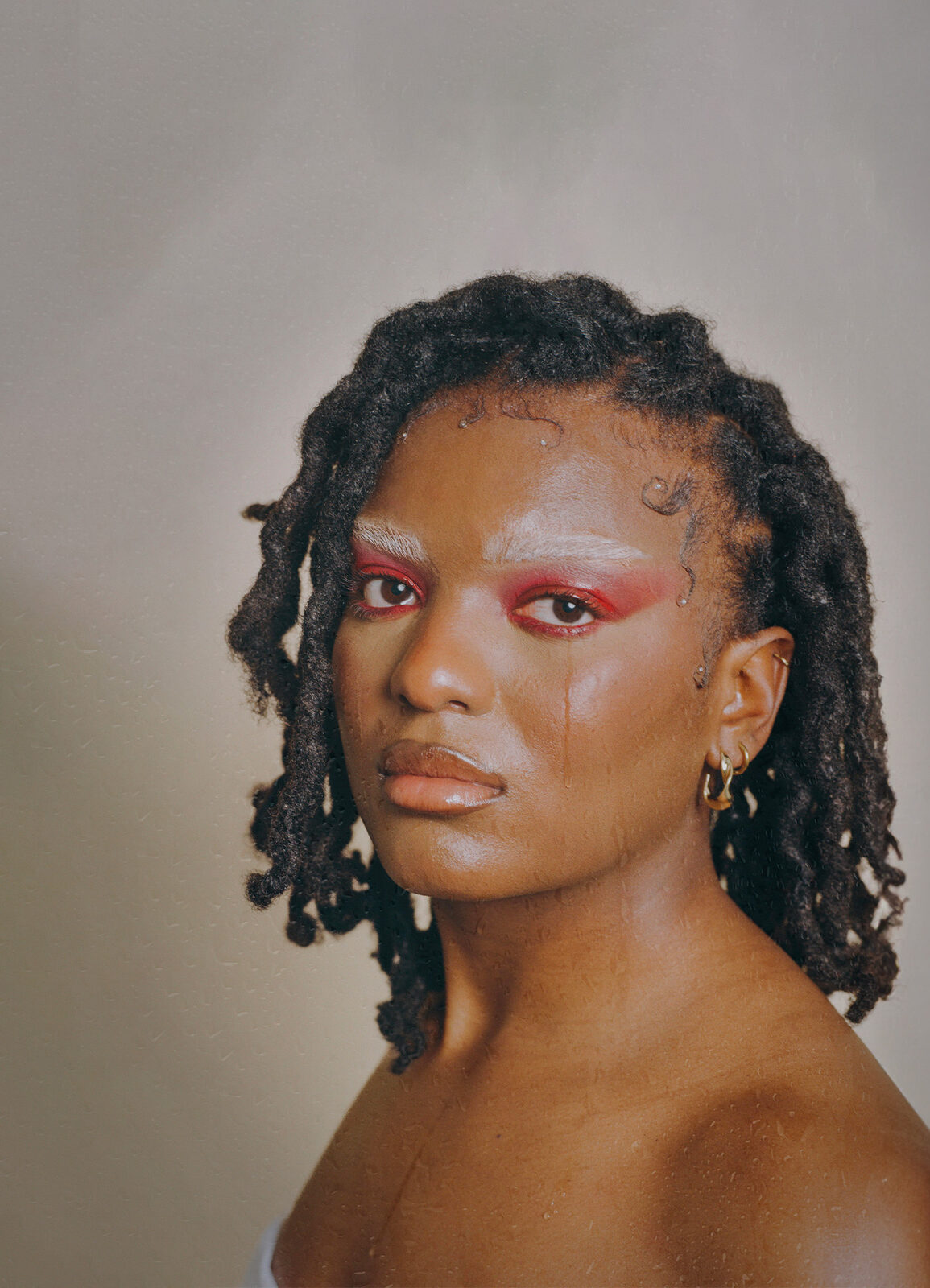 Portrait of a dancer with shoulder-length locs and graphic red-and-white eye make-up looking straight at the camera against a pale grey backdrop