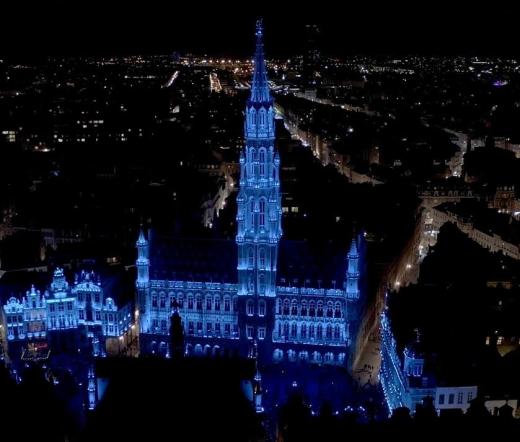 The City Hall tower illuminated in blue at night, seen from above over the rooftops of Brussels city centre.