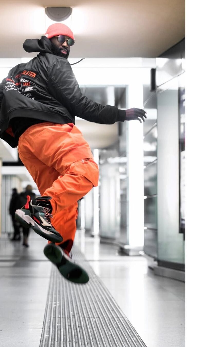 A dancer in bright orange trousers and a black jacket captured mid-jump in a metro station corridor.