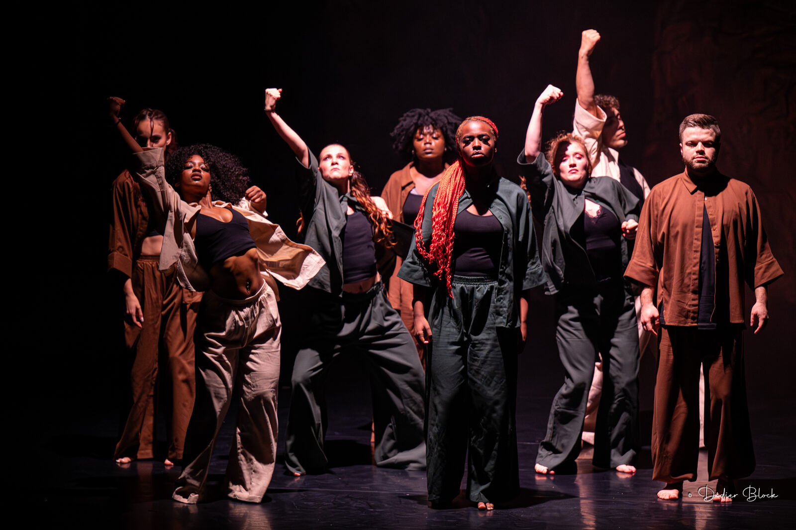 Dancers of the RISE Training Program on a dark stage in earth-toned outfits, several with a fist raised, lit by warm side light.