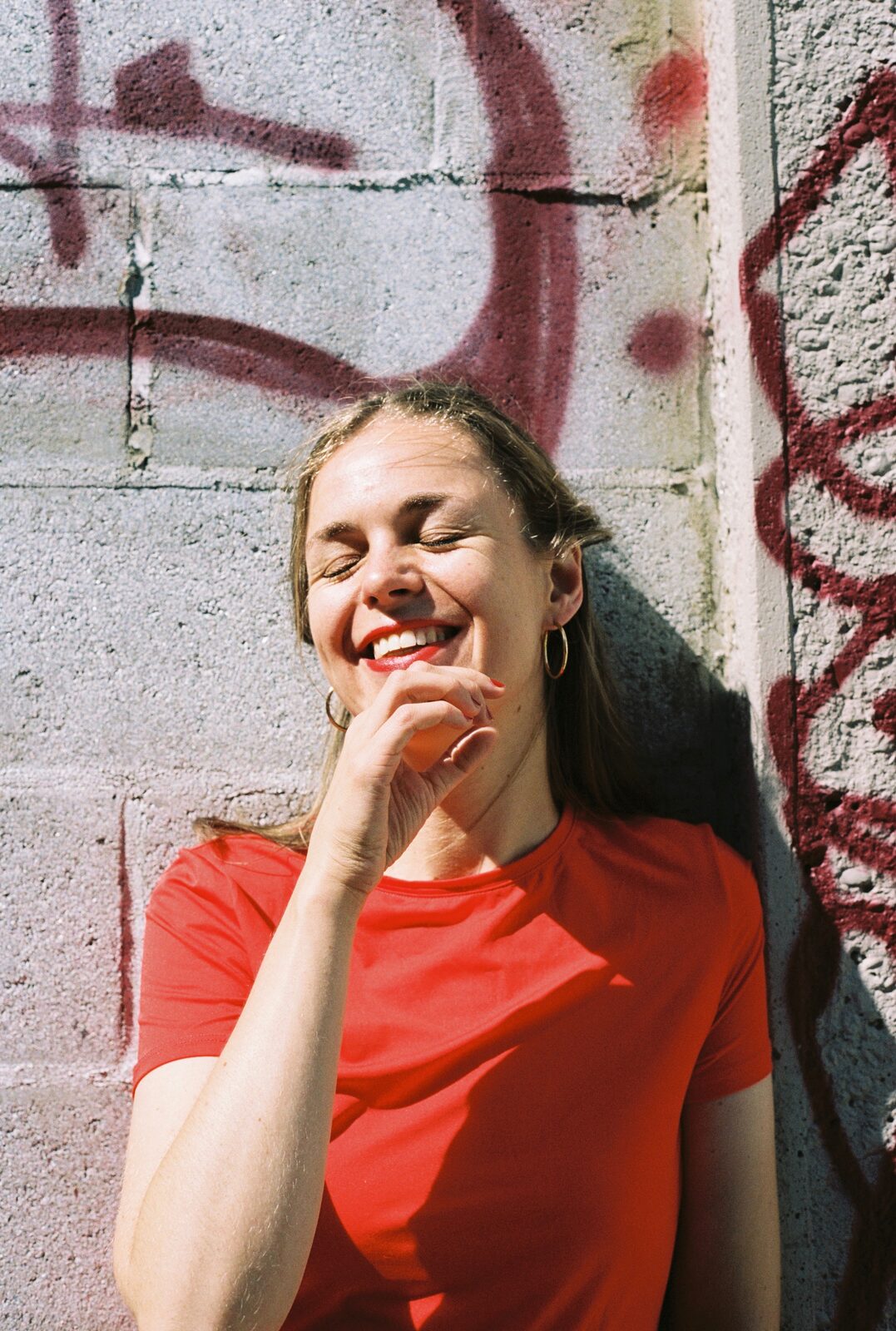 A portrait of a smiling woman in a bright red top laughing in front of a graffiti-covered wall.
