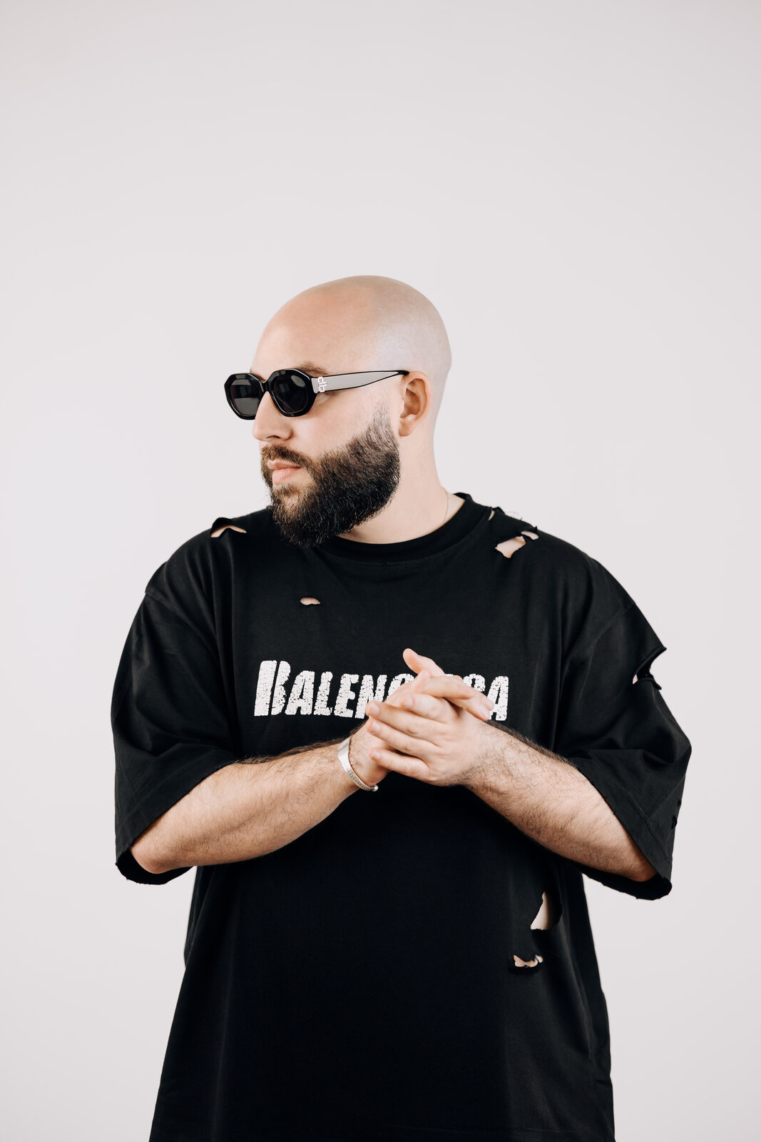 A bearded DJ in dark sunglasses and an oversized distressed black T-shirt standing with his hands clasped against a plain white backdrop