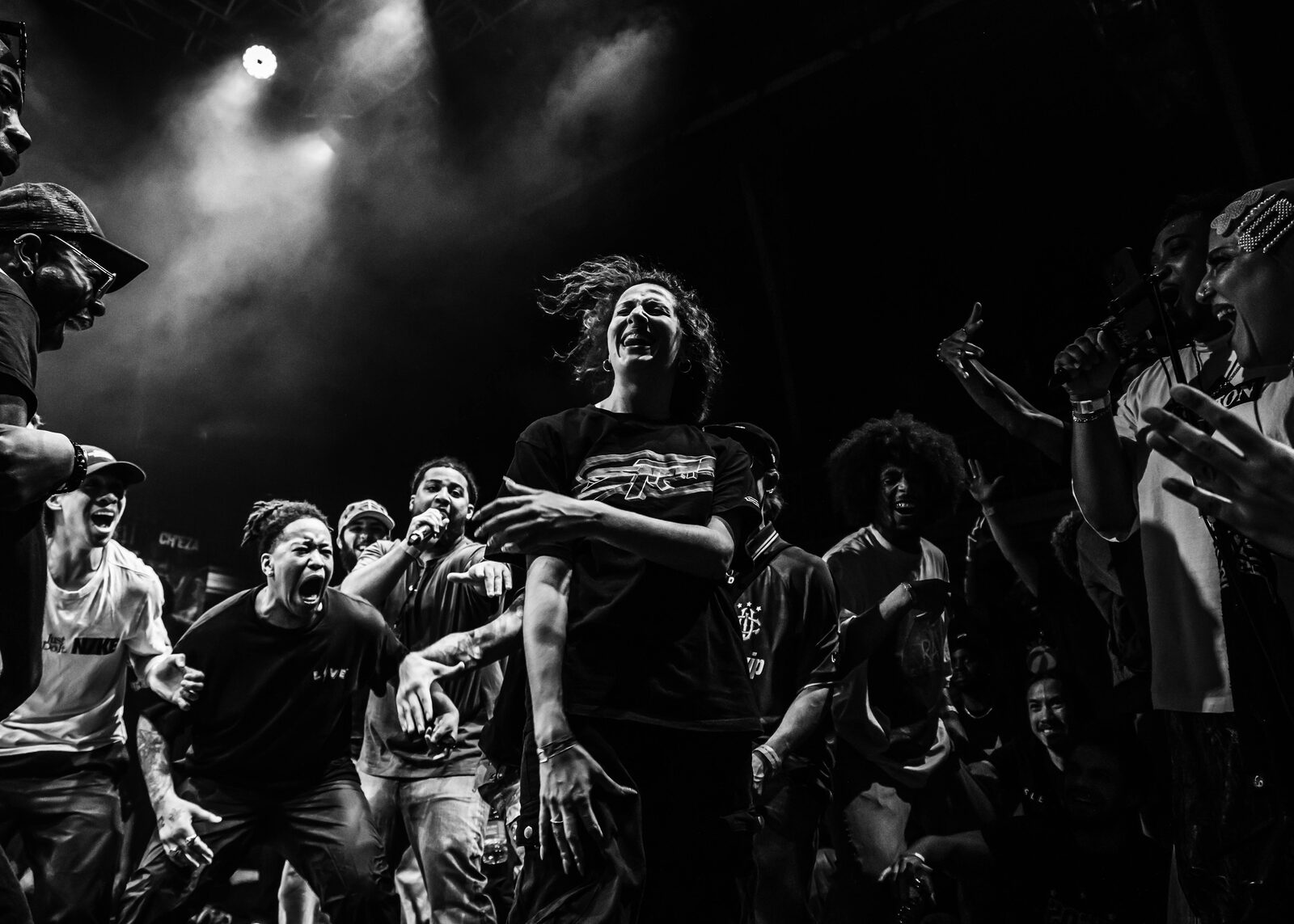 A dramatic black-and-white shot of a dancer mid-battle, surrounded by an energised crowd shouting and cheering under stage lights.