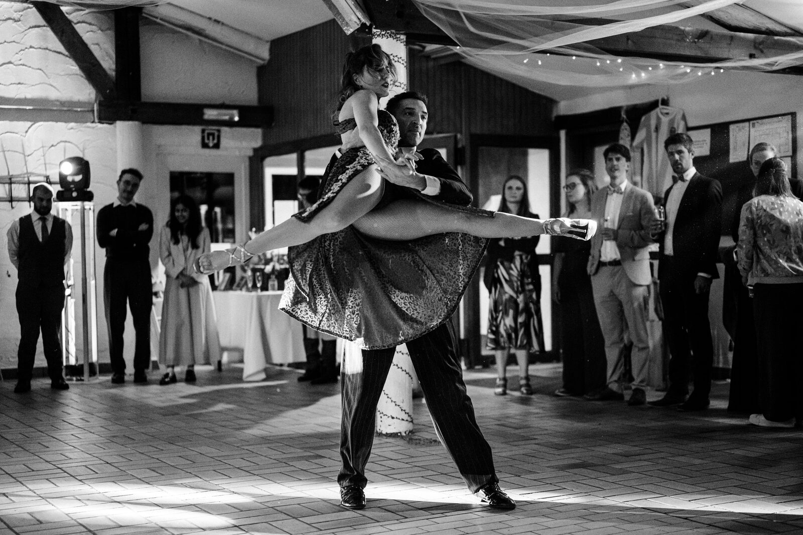 A tango couple performing a dramatic lift on a dancefloor, surrounded by a watching crowd in evening wear.