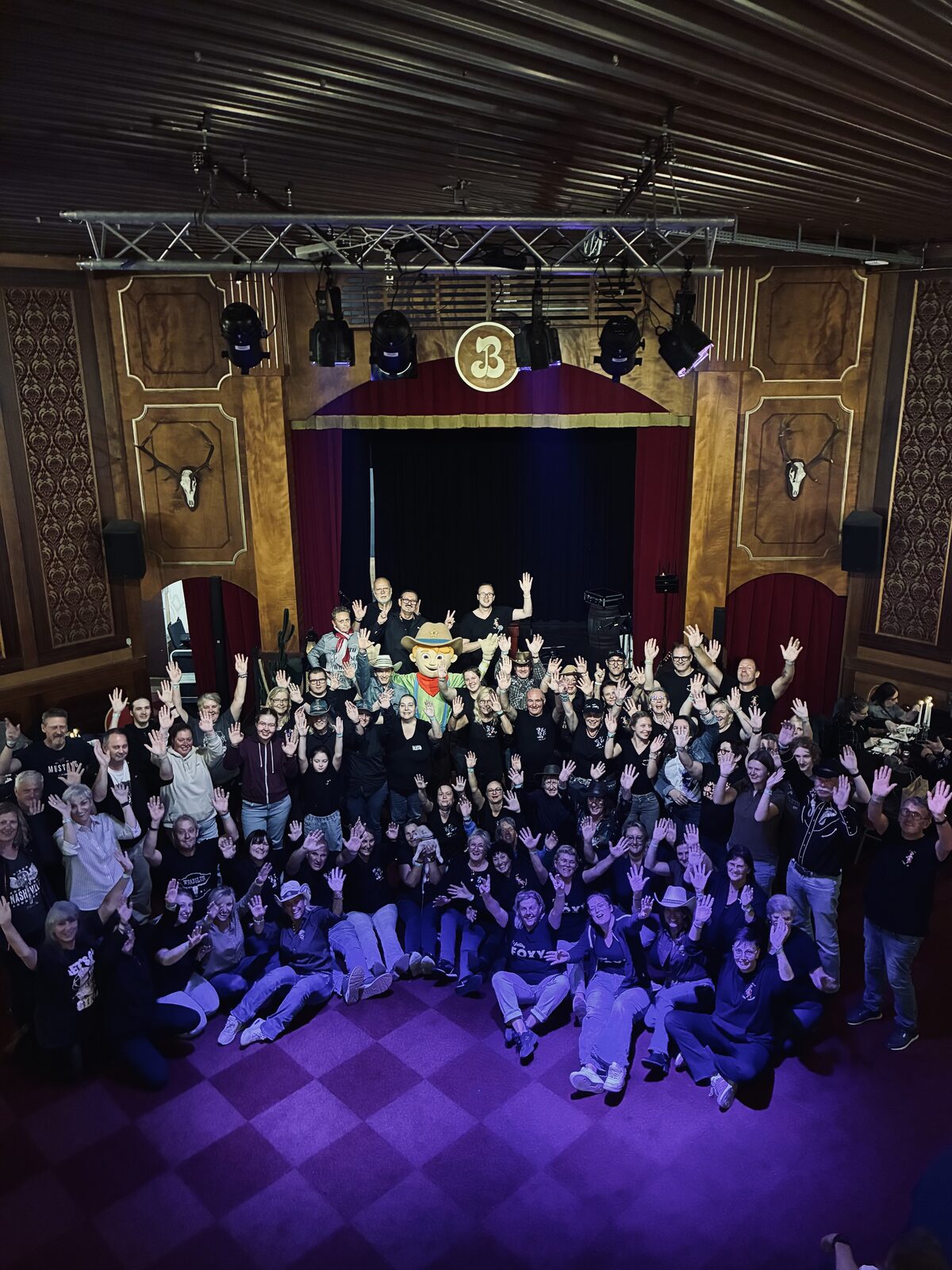 A large group of line dancers in matching western-style outfits posing and waving on a saloon-themed stage.