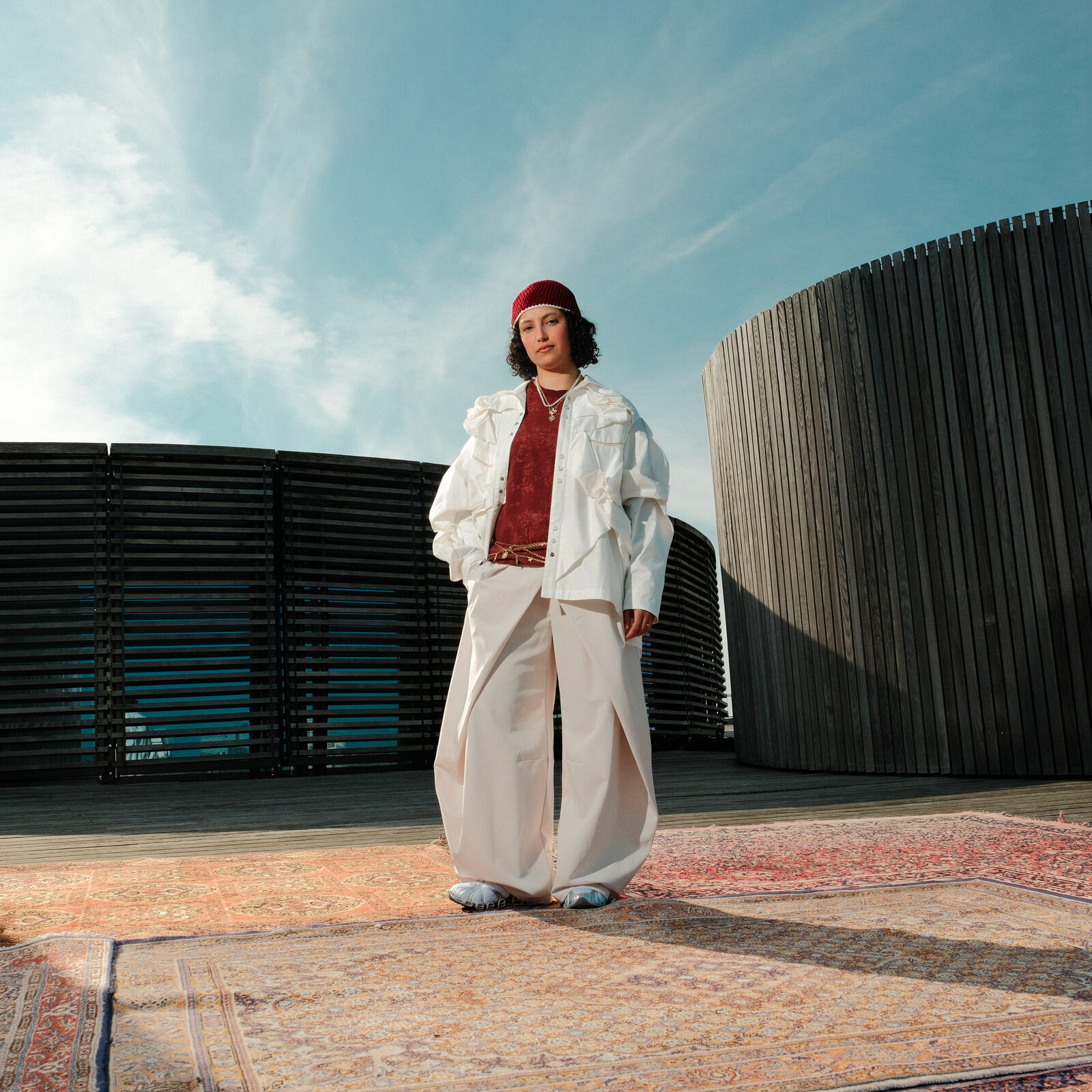 A dancer in a white jacket, wide cream trousers and a red beanie standing on a Persian rug on a rooftop terrace, against slatted dark wood and a blue sky